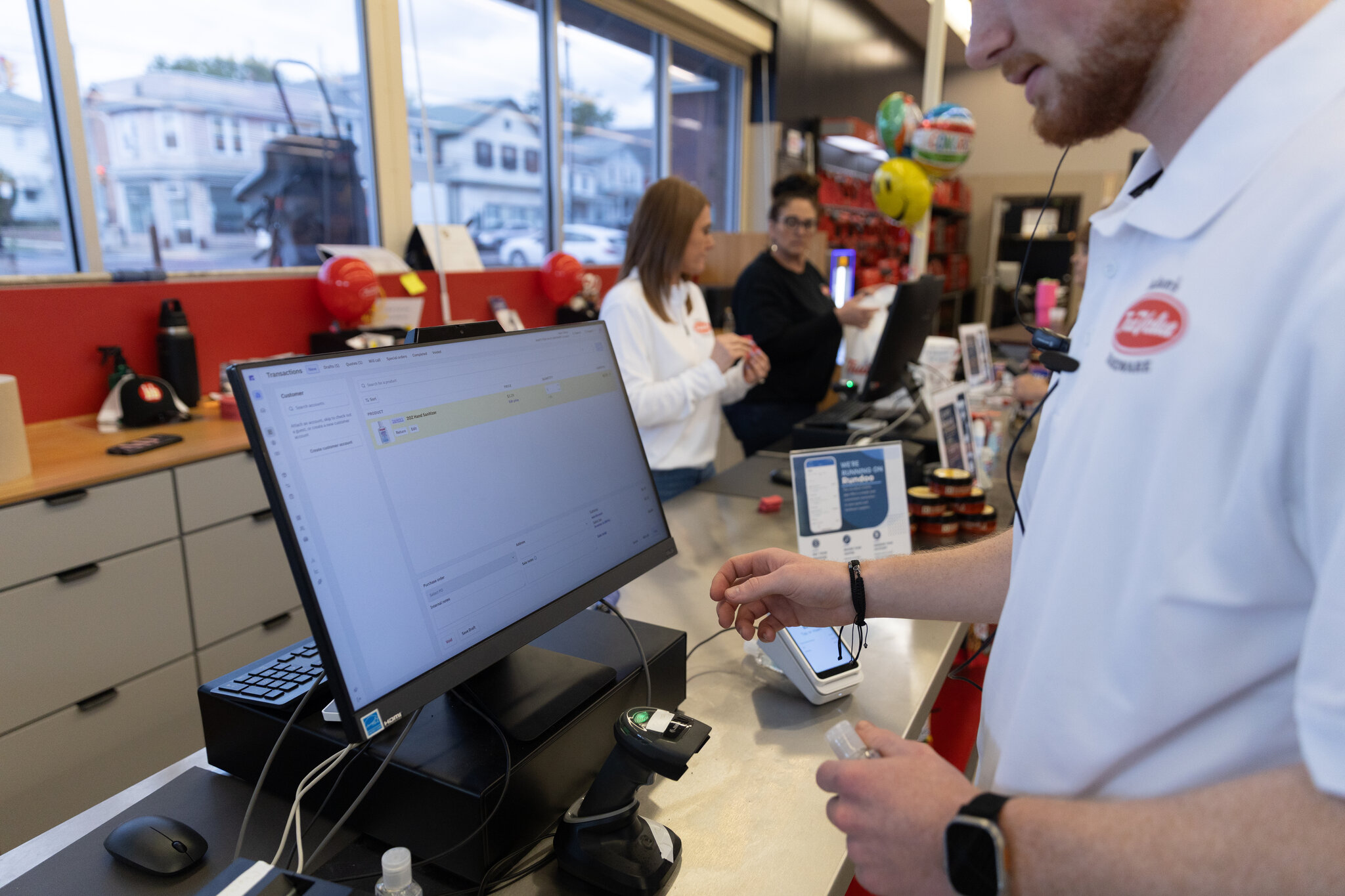 Cashier ringing up a sale on the Rundoo POS at Adam's True Value, with a 'We're running on Rundoo' card on the counter