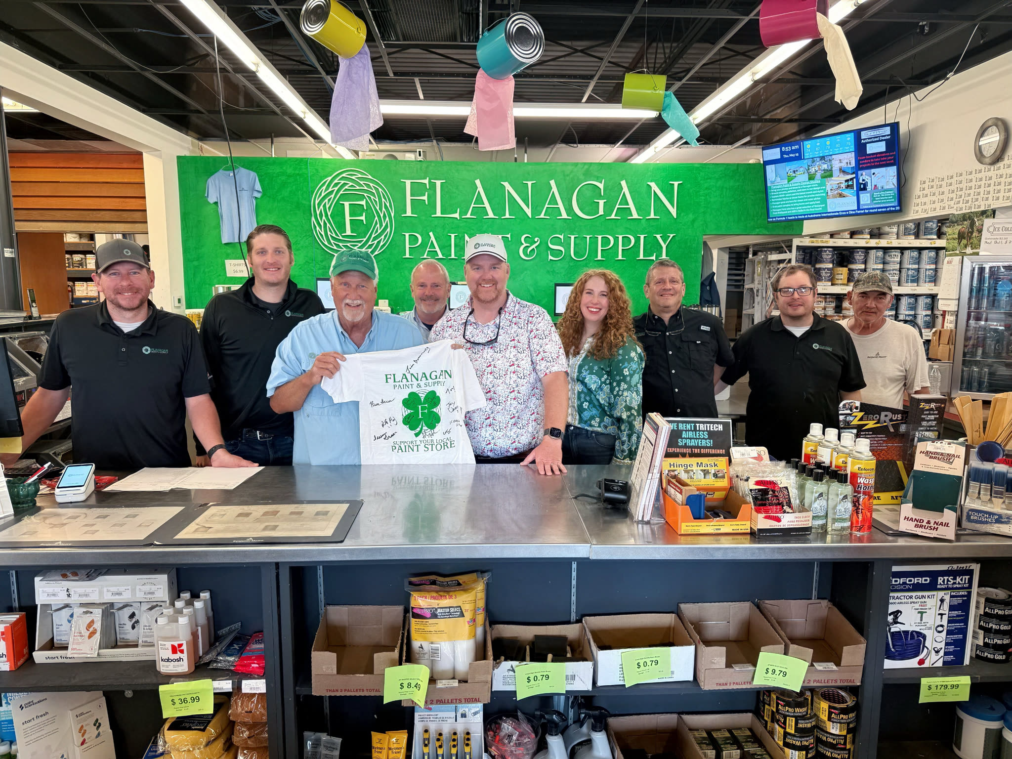 The Flanagan Paint & Supply team behind the counter, holding a signed Rundoo go-live shirt under the store's sign