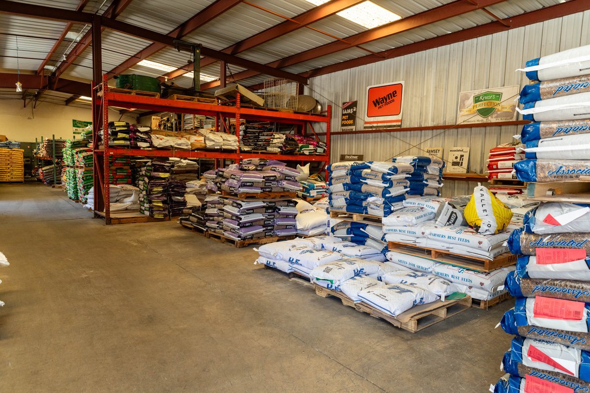Feed bags stacked on pallets and racking in a farm and feed store warehouse, with Purina, Wayne and Farmers Best Feeds signage