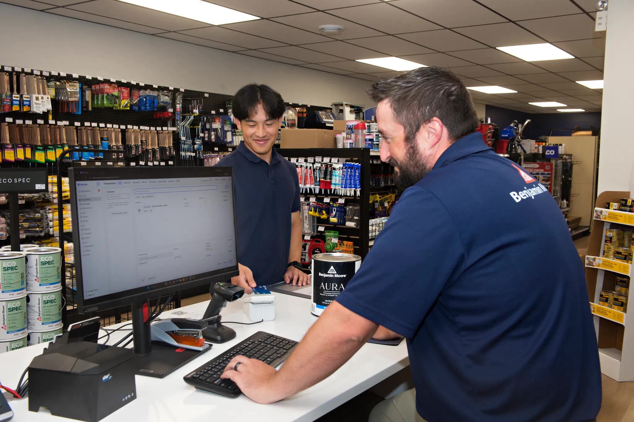 Two people at a paint store counter, one working in Rundoo on the monitor while the other pays by card, with a can of Benjamin Moore Aura on the counter