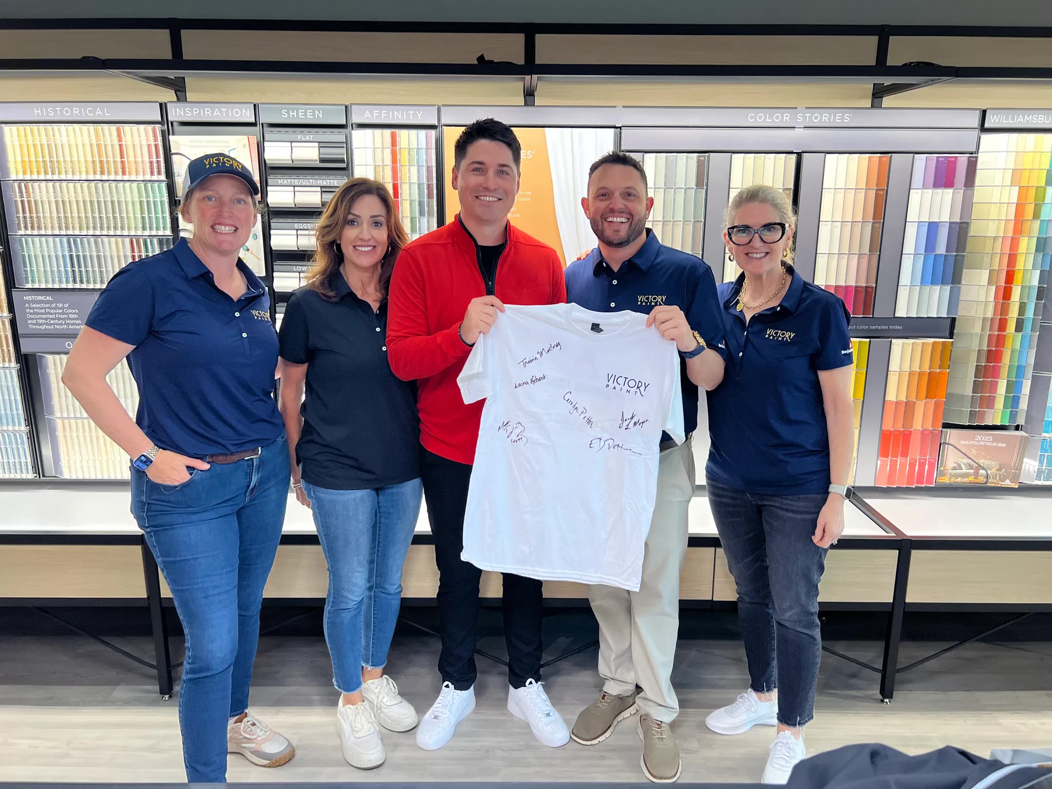 The Victory Paint team holding up their signed Rundoo go-live shirt in front of the Benjamin Moore color displays in their store